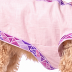 A close-up of a light pink dog coat or sweater with purple and pink geometric patterned trim, shown on a fluffy light tan dog against a white background.