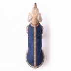 A small light-colored dog viewed from above wearing a navy blue sleeveless pet coat with ornate gold and beige patterned trim along the edges and center front panel, photographed on a plain white background.