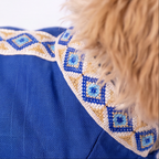 Close-up of a blue pet coat or jacket with a decorative embroidered collar featuring geometric diamond patterns in blue, beige, and white, with a light tan furry animal body visible at the edge.