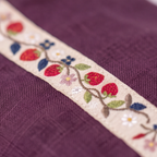 Close-up of a cream-colored embroidered trim featuring a strawberry and floral vine pattern, laid diagonally over a textured purple fabric background.